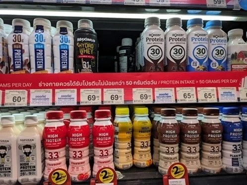 タイのスーパーのプロテインRTD売場。冷蔵棚に各社のボトルが並び、中央の赤い帯に1日50グラム以上のプロテイン摂取を促す表示
